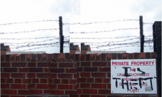 brick wall with barbed wire on top and a sign that has been grafittied to read Private Property is Theft