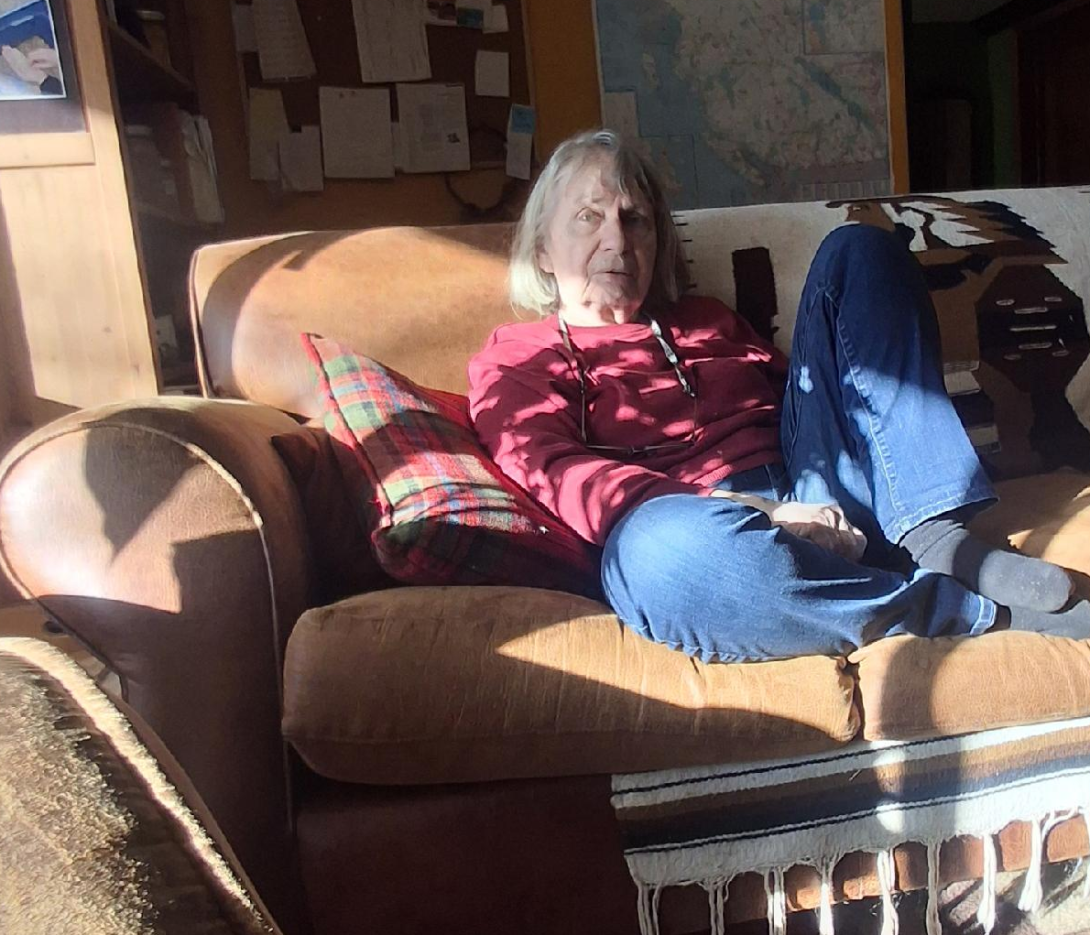 Heather Pritchard in a red sweater, seated on a brown couch with the afternoon sunlight streaming in.